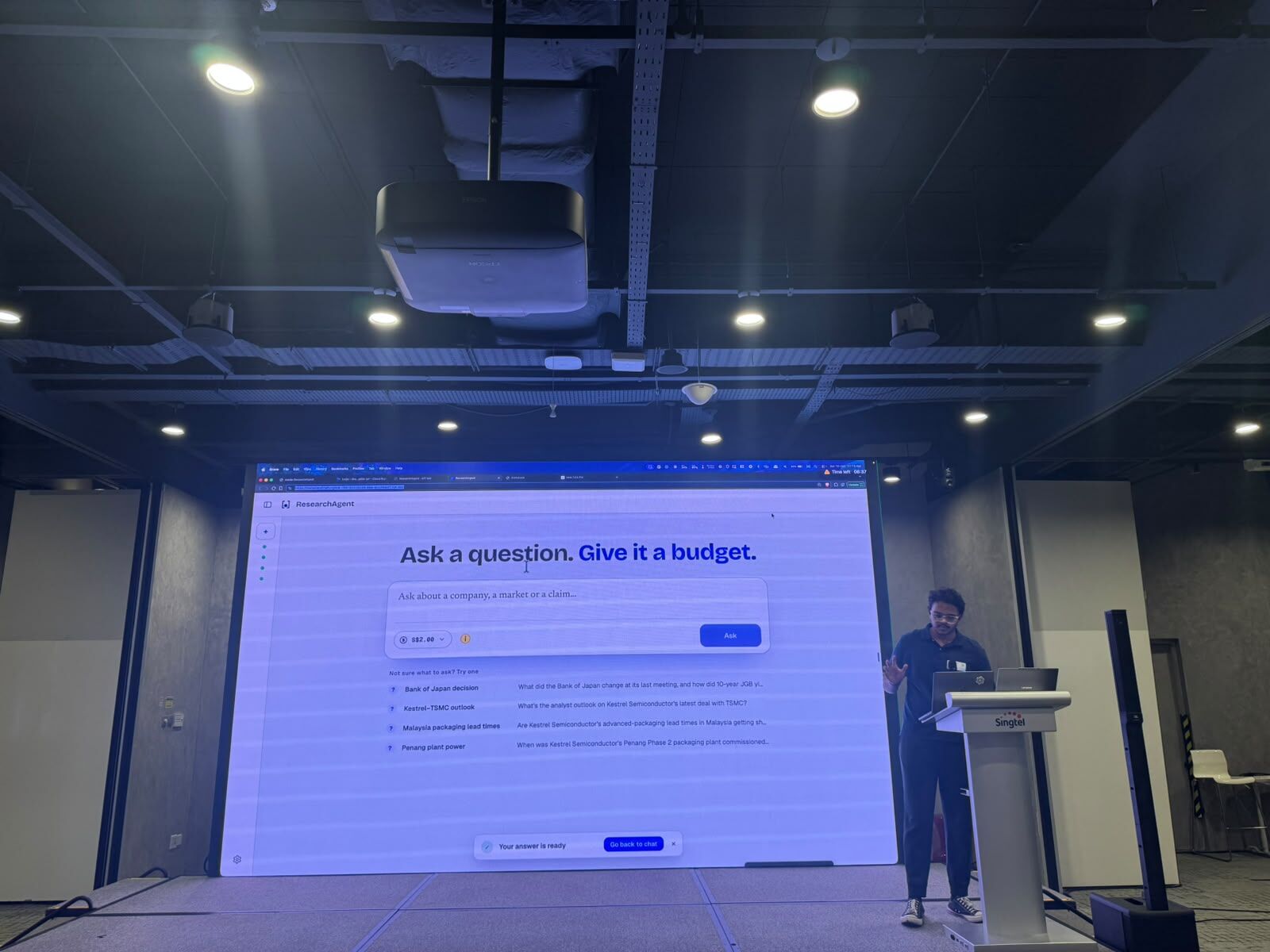 Presenting ResearchAgent on stage at AI Tinkerers Singapore; the screen reads Ask a question. Give it a budget.